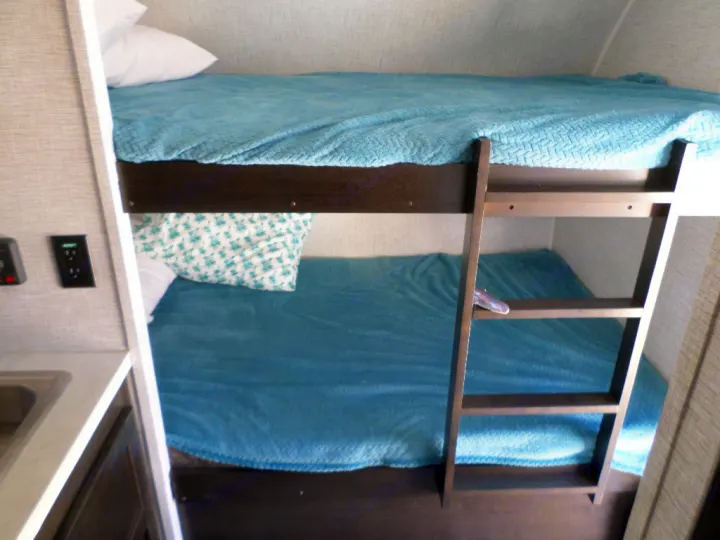 This RV rental features bright bunk beds with blue blankets and a ladder. A compact sink area is visible on the left for comfortable travel.. Braxton Creek Bushwhacker Plus 2022