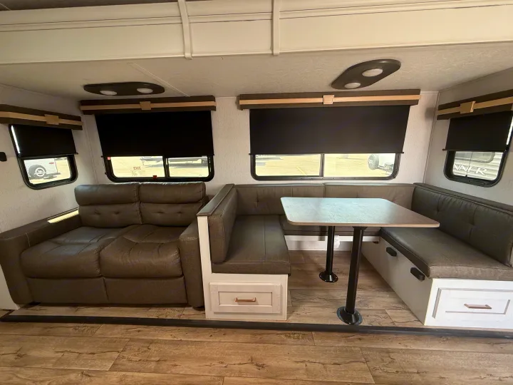 This RV rental interior features a cozy sofa, a U-shaped dinette with a table, and black window blinds, offering a comfortable living area.. Astoria Astoria Trailer 2022