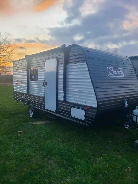 This gray & white Coleman travel trailer, newly sold & parked on grass at sunset, perfectly suits your next RV rental adventure.. Dutchmen Coleman Lantern LT 2021