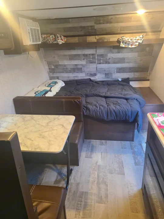 Inside an RV rental: a comfortable bed with gray bedding, a practical dinette with a marble-look table, and an AC unit.. Dutchmen Coleman Lantern LT 2021