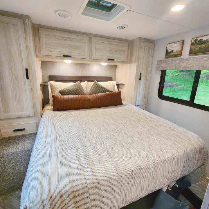 Modern RV rental bedroom. Features light wood cabinetry, a comfy bed with striped comforter, and scenic window views.. Forest River Sunseeker LE 2022