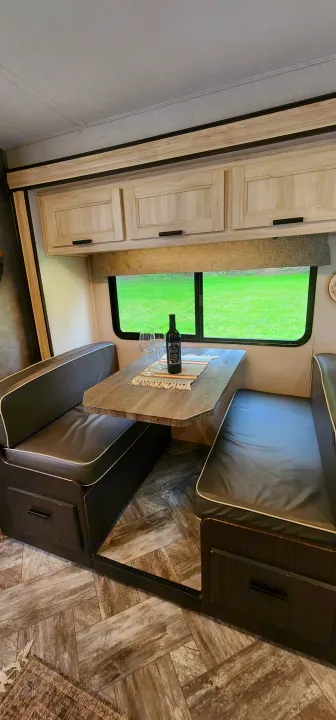 Inside an RV rental: a modern dinette with dark benches, light wood cabinets, and a scenic green view. Wine on table.. Forest River Sunseeker LE 2022