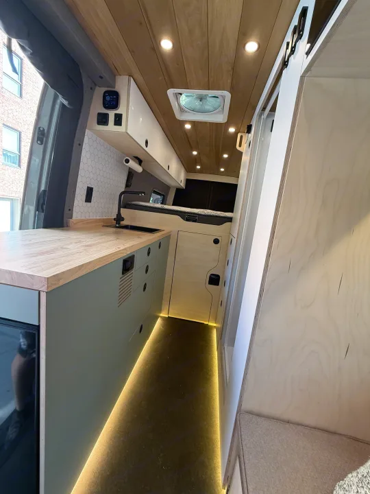 This rv rental features a modern camper van interior with a sleek kitchenette, comfy bed, and wood-paneled ceiling.. Mercedes-Benz sprinter 2024