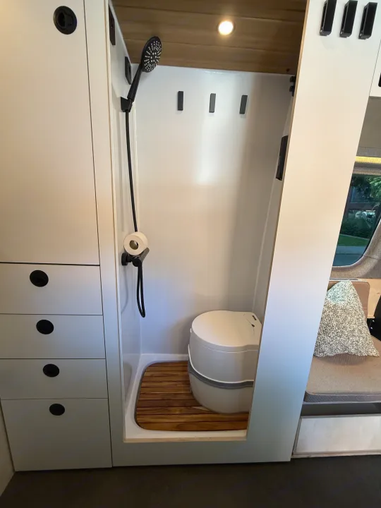 This rv rental features a compact, modern bathroom with a shower, portable toilet, wood slat floor, and storage cabinets.. Mercedes-Benz sprinter 2024