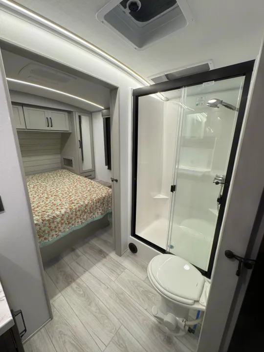 RV rental showing cozy bedroom with floral bedspread beyond a bright, modern bathroom featuring a shower and toilet. Ready for your trip!. Keystone Outback 291BH 2025