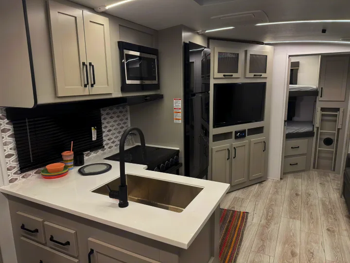 Modern RV rental interior with a bright kitchen featuring white counters, stove, sink, microwave, TV, and cozy bunk beds.. Keystone Outback 291BH 2025