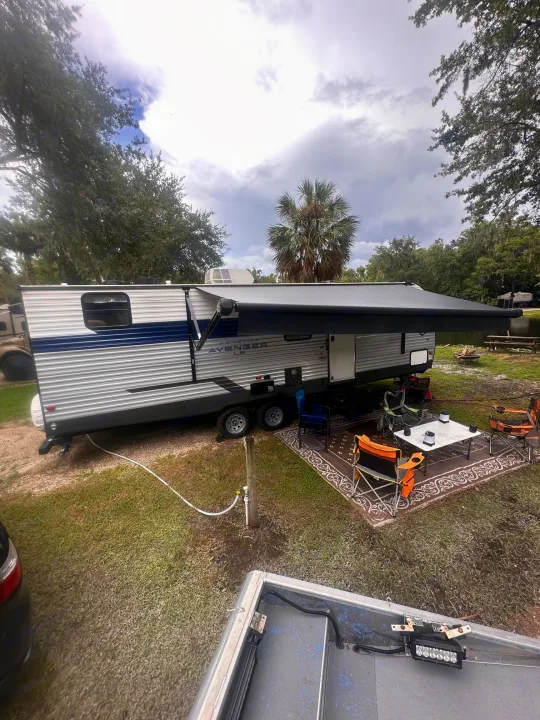 RV rental travel trailer campsite with extended awning, outdoor living area, and boat by the water.. Forest River Avenger LE 2022