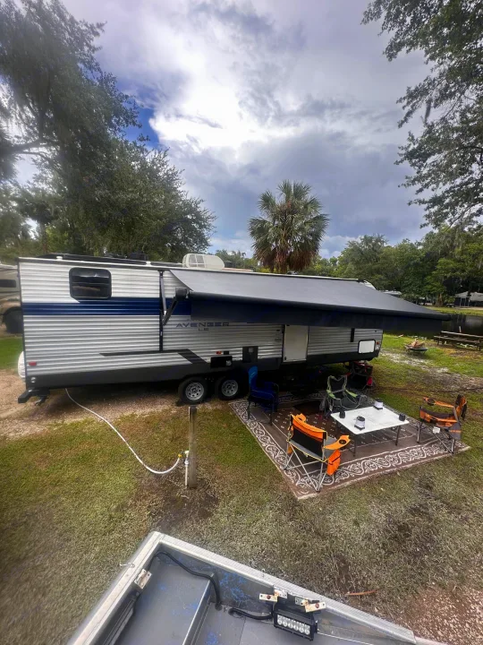 RV rental camping setup: travel trailer with extended awning, outdoor rug, table, and chairs under cloudy skies.. Forest River Avenger LE 2022