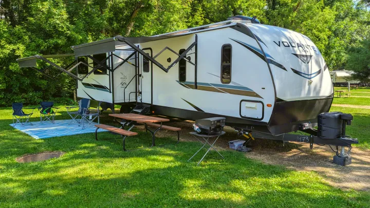 RV rental setup at a sunny campground. A white travel trailer with awning extended, picnic table, chairs, and grill on grass.. Crossroads RV Volante 2022
