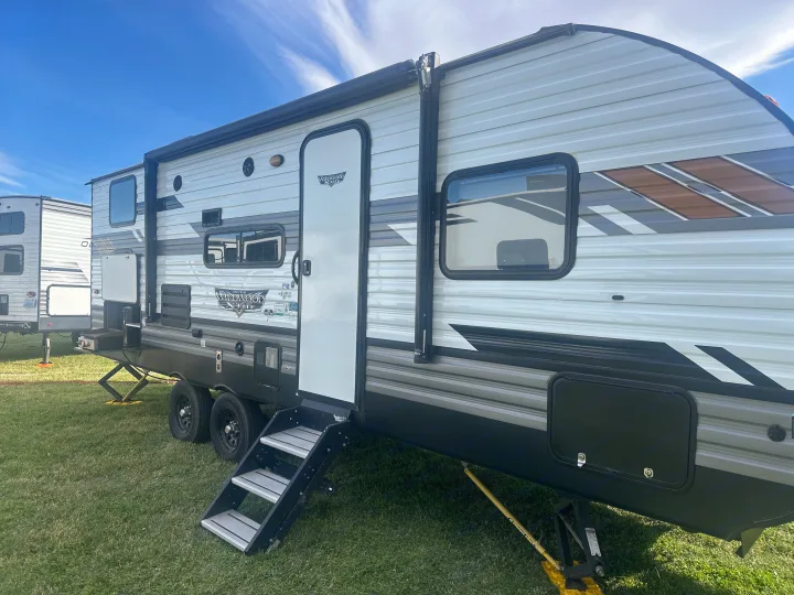 An RV rental setup: a large white/grey travel trailer with steps and leveling jacks on green grass, ready for camping.. Forest River Wildwood X-Lite 2022