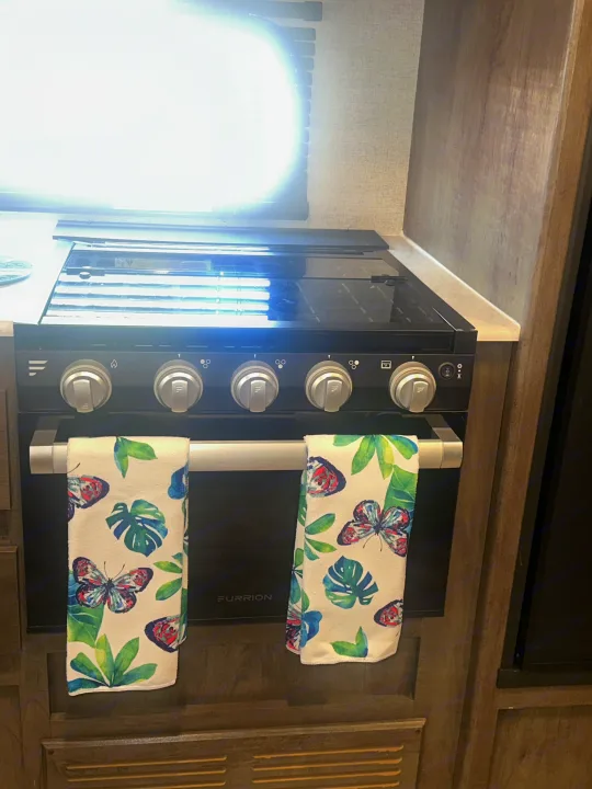 RV rental kitchen showing a black Furrion stove with control knobs and butterfly towels on the oven handle, next to a bright window.. Forest River Wildwood X-Lite 2022