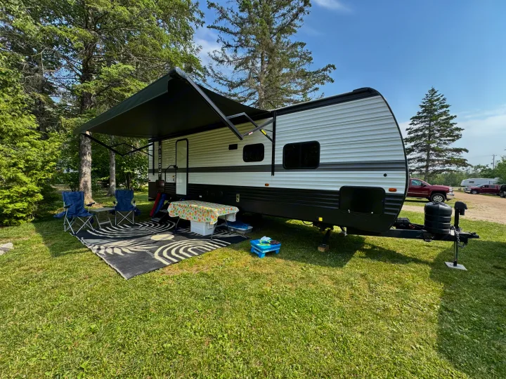 Serene RV rental site: A white travel trailer with an extended awning, outdoor chairs, and a table on green grass.. Keystone RV Hideout 2024