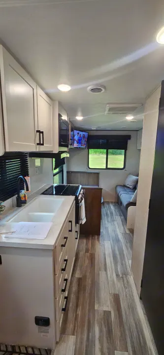 Modern RV rental interior: a bright kitchen with white cabinets, sink, and stove, plus a living area with a TV.. Jayco Jay Flight SLX 210 QB 2025