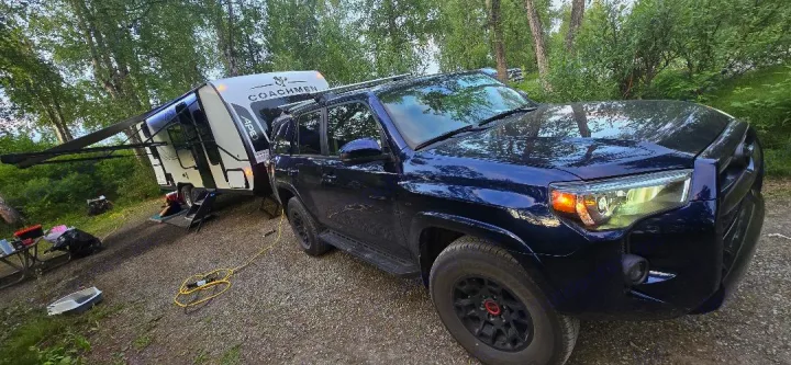 Dark blue SUV towing a white Coachmen RV rental at a wooded campsite, ready for an outdoor adventure.. Coachmen Apex 2025