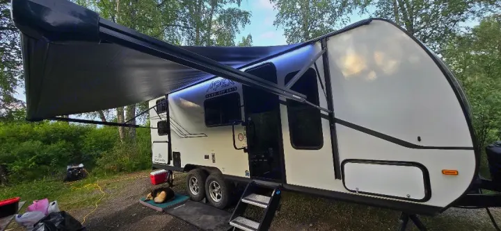 An RV rental, Apex Nano-Off Grid model, is set up at a leafy campsite with its awning extended. A dog naps nearby.. Coachmen Apex 2025