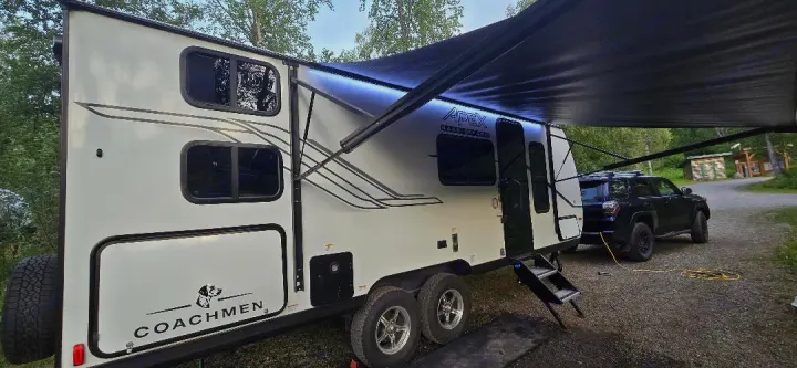 A white Coachmen Apex travel trailer with its awning extended at a woodsy site, parked with an SUV. Ideal RV rental.. Coachmen Apex 2025