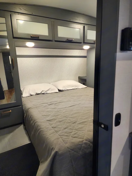 Modern RV rental bedroom: cozy grey bed with white pillows, geometric headboard, and sleek overhead cabinets with lights.. Keystone RV Bullet Ultra Lite 2023
