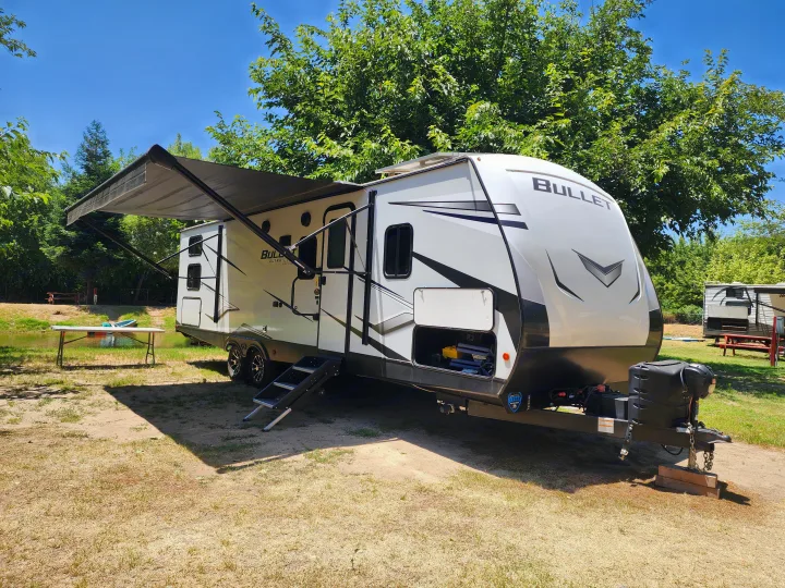 Enjoy an RV rental! This modern travel trailer is perfectly set up at a lakeside campsite under a blue sky.. Keystone RV Bullet Ultra Lite 2023