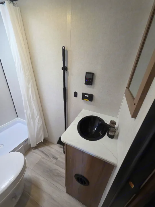 RV rental bathroom features a white shower, toilet, and a vanity with a sleek black sink. Wall controls & wood-look floor. Compact & functional.. Keystone Coleman 2025
