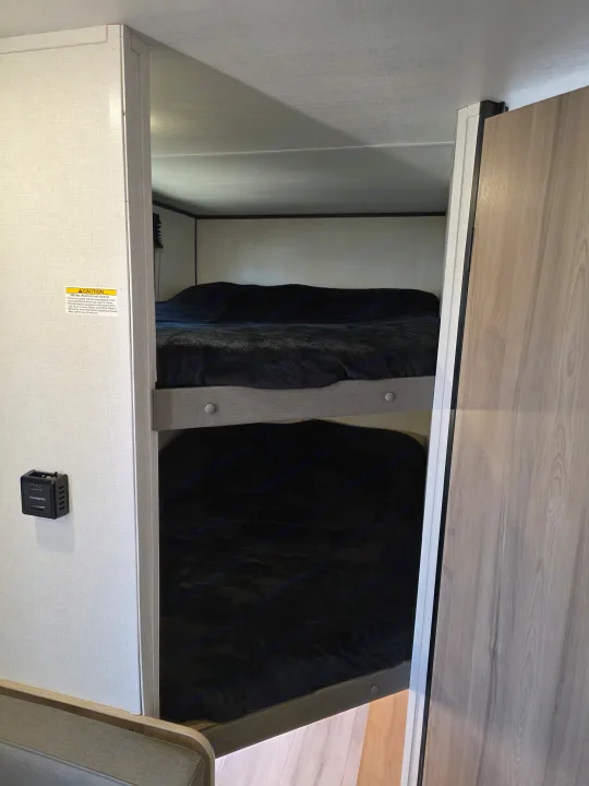 RV rental bunk beds are visible, featuring two cozy bunks with black blankets, a thermostat, and a bed safety warning.. Keystone Coleman 2025
