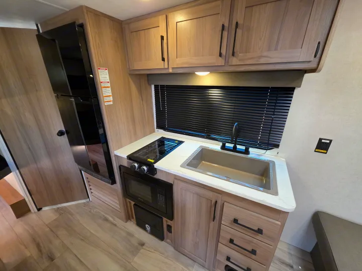 This modern RV rental kitchen features light wood cabinetry, black appliances (fridge, stove, microwave), and a sink. Compact and functional.. Keystone Coleman 2025
