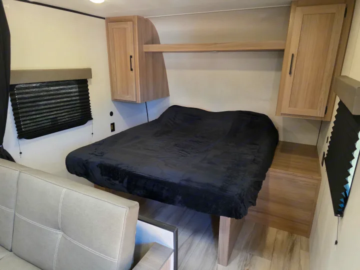 This RV rental features a cozy bedroom with a black blanket on the bed, overhead cabinets, a window, and a sofa.. Keystone Coleman 2025