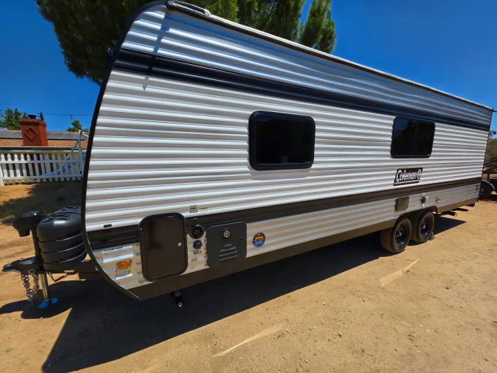 A sleek Coleman travel trailer, ideal for rv rental, parked on a sunny dirt lot with trees and a house in the background.. Keystone Coleman 2025