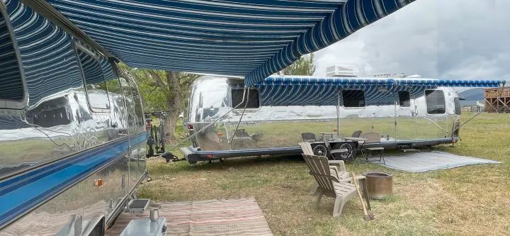 Two shiny Airstream RVs, possibly for rv rental, parked with striped awnings extended. Outdoor setup includes chairs and a fire pit.. Airstream Excella 1990