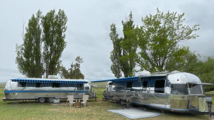Two shiny Airstream RVs with blue awnings extended, set up for outdoor camping in a grassy field. Ideal for an RV rental.. Airstream Excella 1990