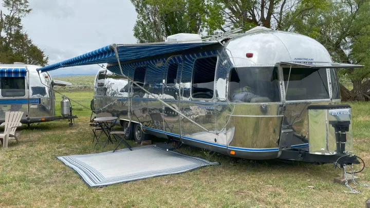 Polished Airstream RV rental with an extended blue awning and outdoor rug on a grassy site, perfect for camping.. Airstream Excella 1990