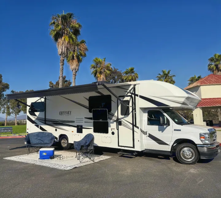 This white Odyssey RV rental is set up for camping with an extended awning, chairs, and cooler under palm trees.. Entegra Coach Odyssey 2022