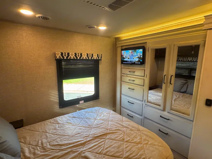 Inside a modern RV rental bedroom, featuring a light-colored bed, window, TV, and mirrored closet storage.. Entegra Coach Odyssey 2022