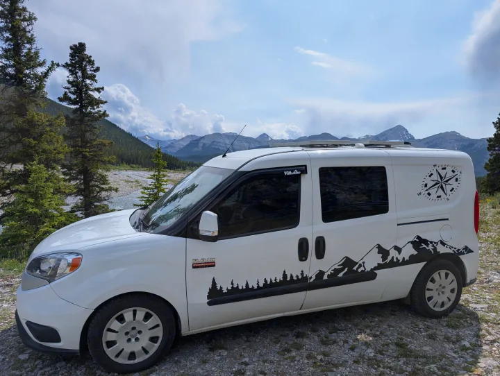 White Ram ProMaster City rv rental with mountain/forest decals, parked by a scenic river & mountains under a cloudy sky. Perfect for van life.. Dodge Sprinter Van 2015