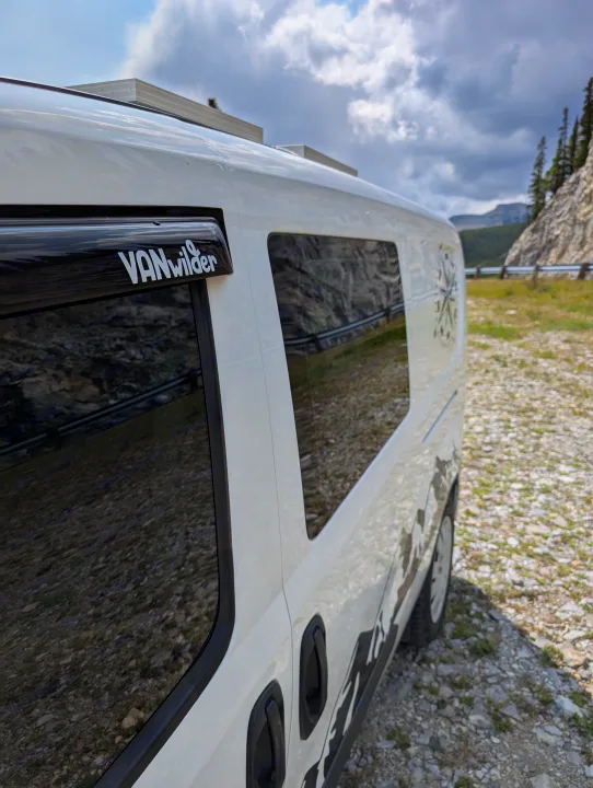 Side view of a white 'VANwilder' campervan, an ideal RV rental, parked on gravel with mountains and cloudy sky.. Dodge Sprinter Van 2015