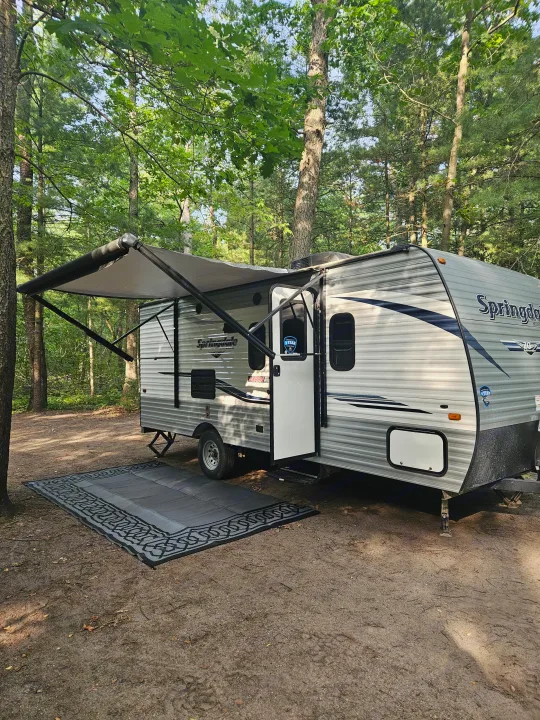 RV rental parked in a peaceful wooded campground with its awning extended and a large outdoor mat laid out.. Keystone RV Springdale 2019