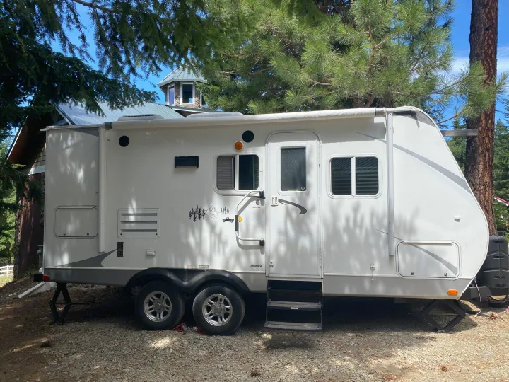 White RV rental travel trailer parked under tall trees, with a unique octagonal building visible in the background.. Cruiser RV Fun Finder X 2011