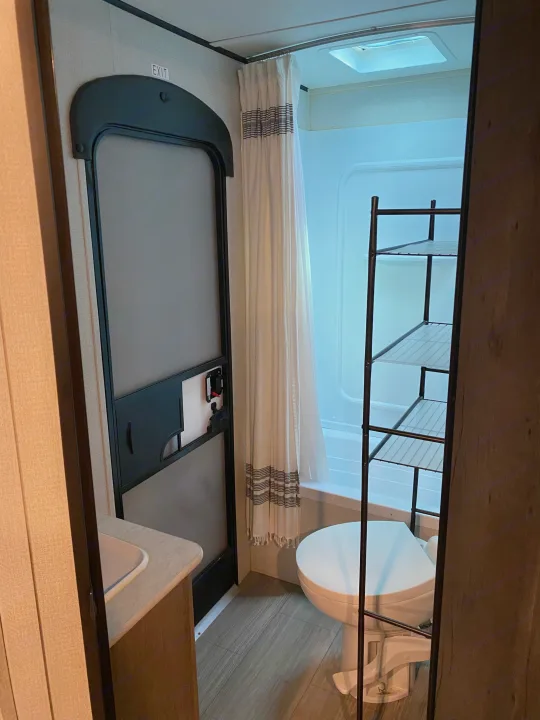 Compact RV rental bathroom showcasing a shower with curtain, toilet, sink, and shelving unit. An exit door is visible.. Keystone RV Hideout 2021