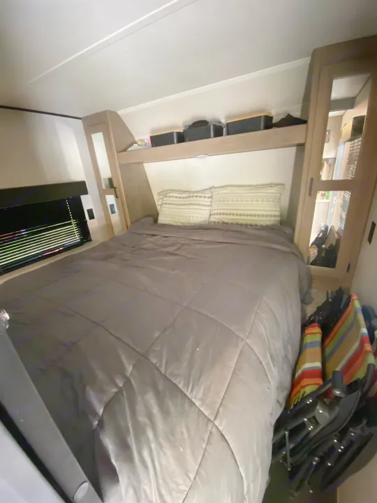 RV rental bedroom: gray bed, overhead storage shelf with boxes, mirrored wardrobe, and window with blinds.. Keystone RV Hideout 2021