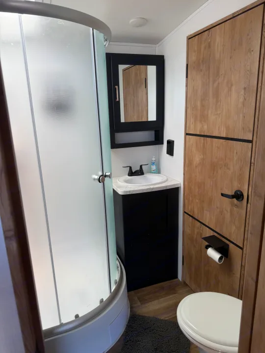 Inside an RV rental bathroom: features a curved frosted shower, black sink vanity, mirrored cabinet, and a toilet with wood accents.. Keystone RV Outback 2022