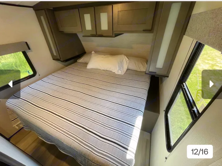 Cozy RV rental bedroom with a striped bed, white pillows, overhead cabinets, and windows showing green outdoor scenery.. Cruiser RV MPG Ultra Lite 2021