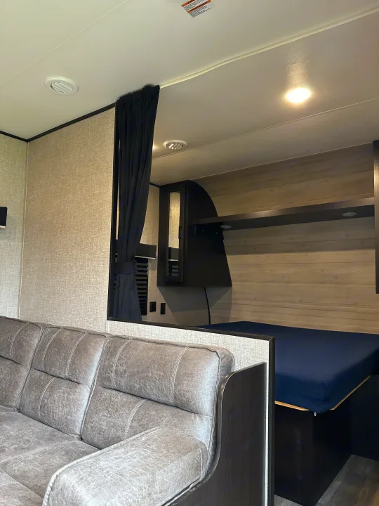 Modern RV rental interior: a comfy gray sofa, dividing wall, and private bedroom with a dark blue bed & wood paneling.. Jayco Jay Flight SLX 2022