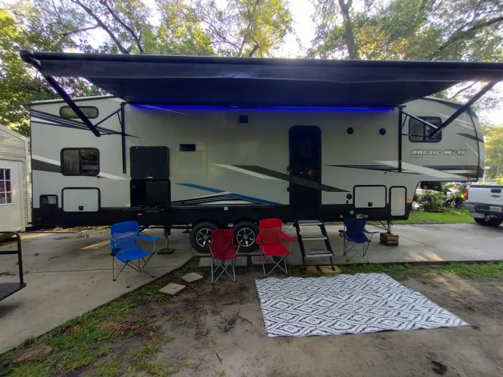 An RV rental with an extended awning and blue lights, set up at a campsite with chairs and a mat for outdoor living.. Forest River Cherokee Arctic Wolf 2021
