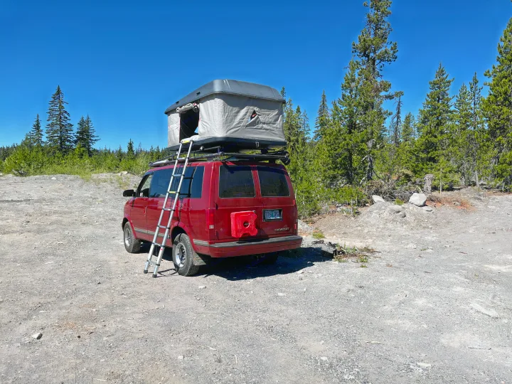 Red RV rental camper van with a rooftop tent and ladder, parked on a sunny gravel path in a forest.. Chevrolet Astro Van 1997