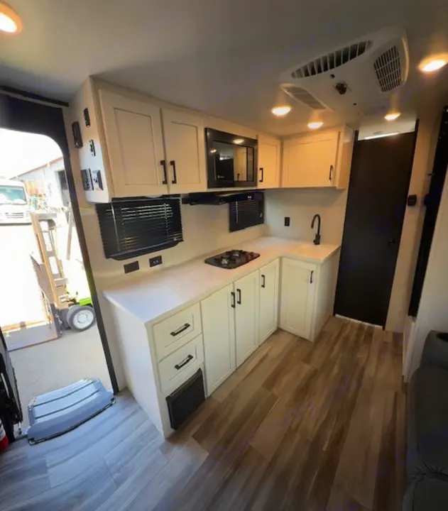 Modern RV rental kitchen interior featuring white cabinets, black appliances, and a wood-look floor. View toward open rear ramp.. JAYCO Jay Flight 2025