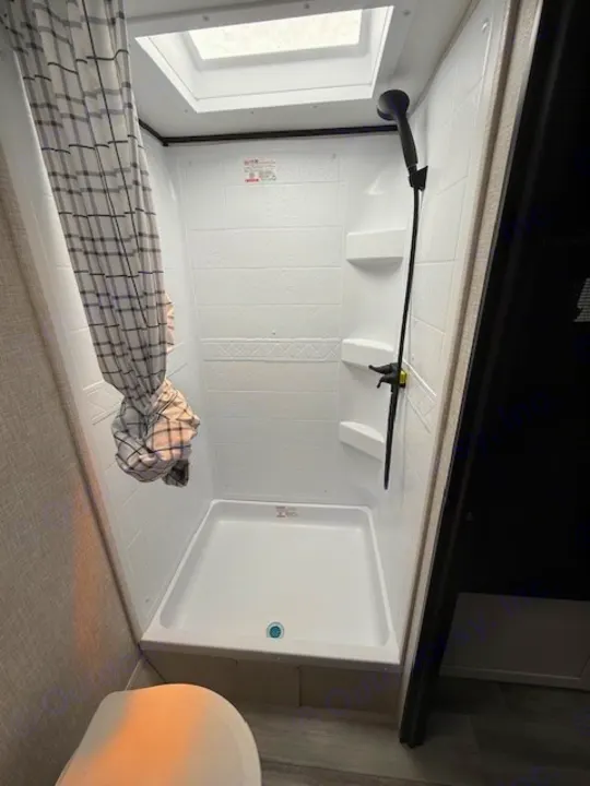 This RV rental bathroom boasts a clean, compact shower with a skylight, black fixtures, and a plaid curtain.. JAYCO Jay Flight 2025