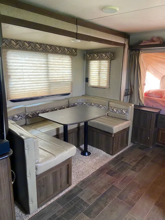 Inside a modern RV rental: a U-shaped dinette with light upholstered seating, dark table, and window blinds, plus a cozy bed area.. Keystone RV Bullet Crossfire 2018