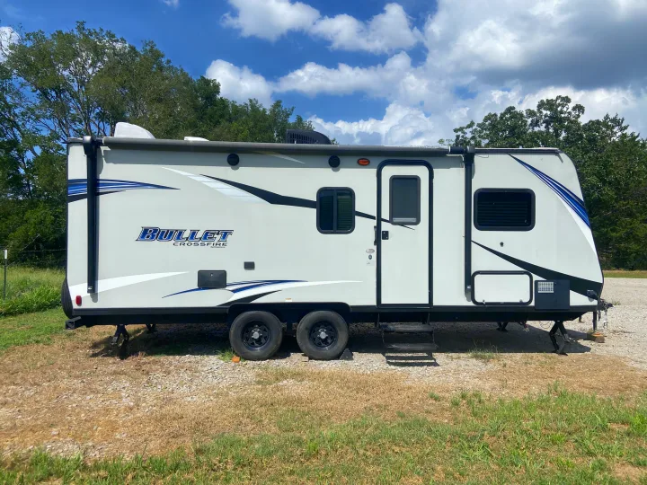 Grey Bullet Crossfire travel trailer, ready for your next rv rental. Parked outdoors on gravel under a sunny, cloudy sky.. Keystone RV Bullet Crossfire 2018