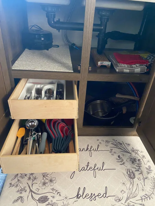 RV rental kitchen under-sink storage showing pipes, a hot water unit, towels, and organized pull-out drawers with utensils.. Keystone RV Bullet Crossfire 2018