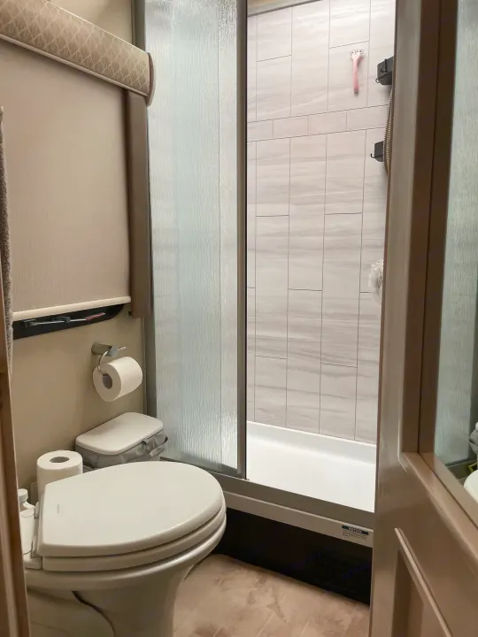 Inside an RV rental, a clean bathroom features a toilet, a shower with modern tiled walls and a textured glass door.. Renegade Custom Coaches Vienna 2021