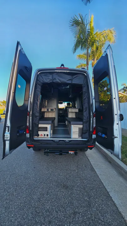 Explore this bright rv rental! Rear doors open on a silver camper van, revealing a custom interior with beds and a kitchenette.. Airstream Interstate 2024
